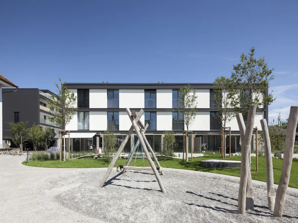 Moderne Wohnanlage mit weißer und dunkler Fassade, umgeben von einer großzügigen Grünfläche und einem Spielplatz mit Holzstrukturen im Vordergrund.