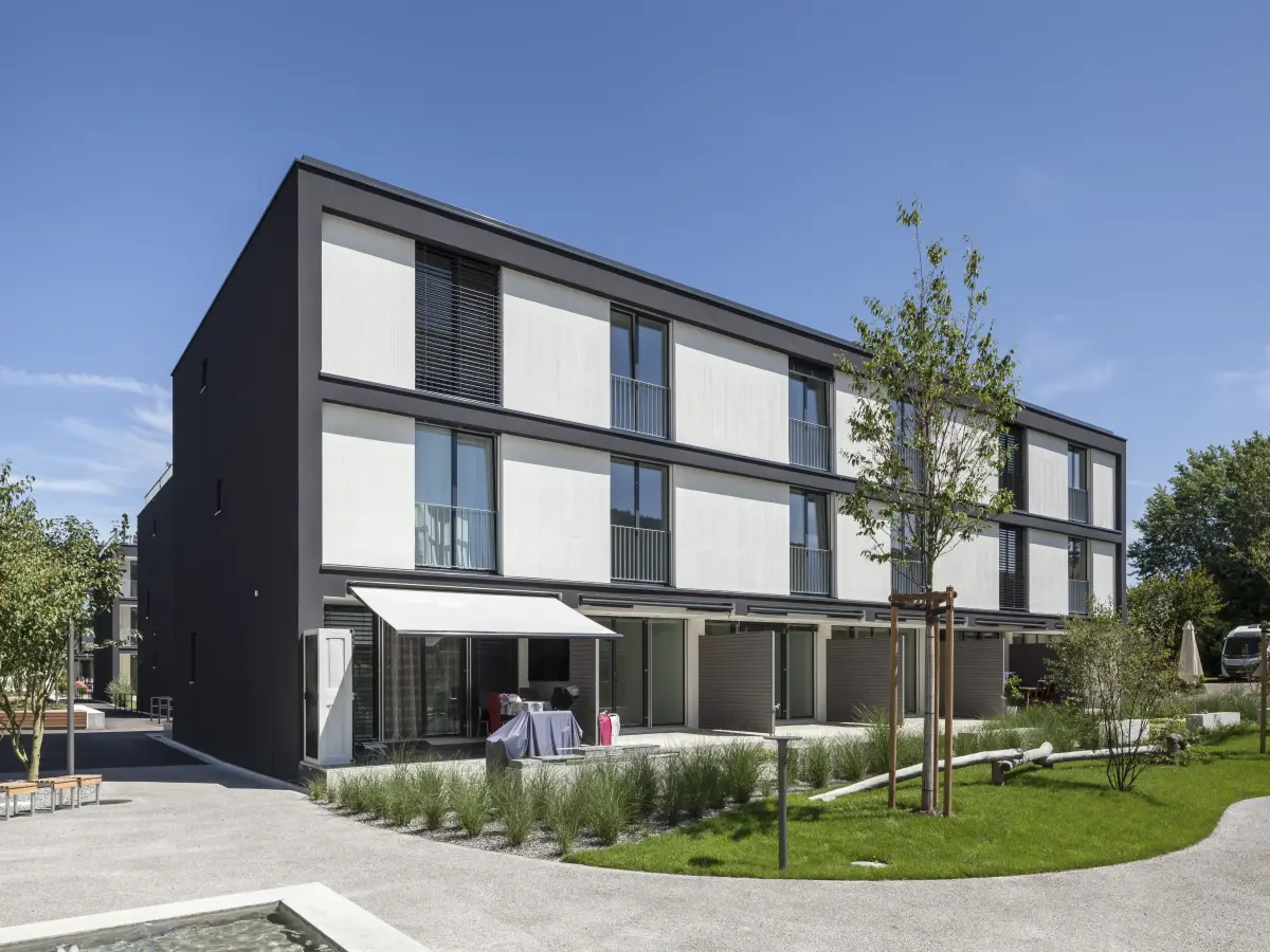 Modernes dreistöckiges Wohngebäude mit dunkler Rahmenfassade, hellen Paneelen und großzügiger Grünanlage im Vordergrund bei strahlend blauem Himmel.