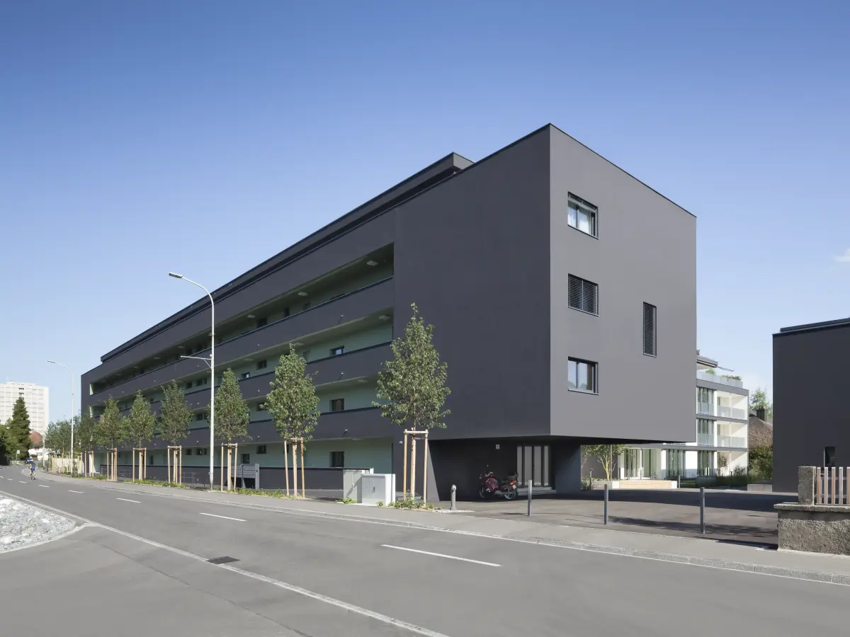 Modernes Wohngebäude mit dunkler Fassade und horizontalen Balkonen, gesäumt von jungen Bäumen entlang einer ruhigen Straße bei klarem Himmel.