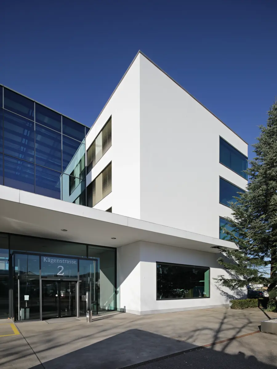 Eingangsbereich eines modernen Gebäudes mit weißer Fassade, Glasfront und der Adresse "Kägenstrasse 2", flankiert von einem Baum, bei strahlend blauem Himmel.