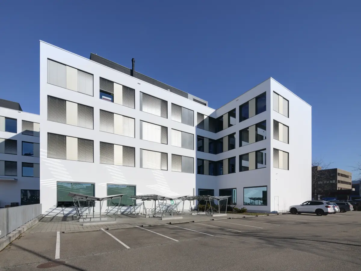 Modernes, weißes Gebäude mit großen Fenstern, umgeben von einem Parkplatz mit Fahrradständern, bei klarem, sonnigem Himmel.
