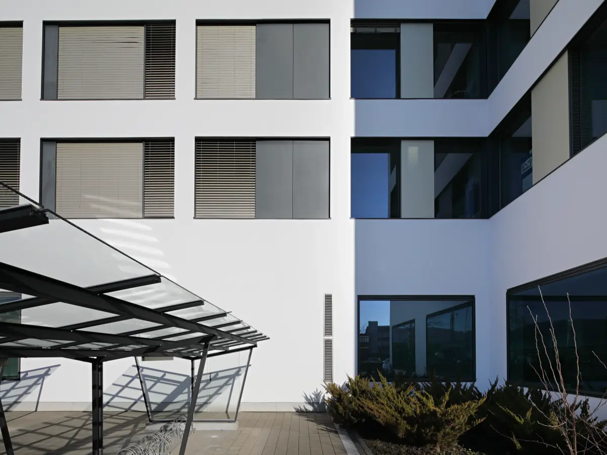 Detailansicht eines modernen, weißen Gebäudes mit Fenstern und Außenverschattungen sowie einem Glasdach im Vordergrund.