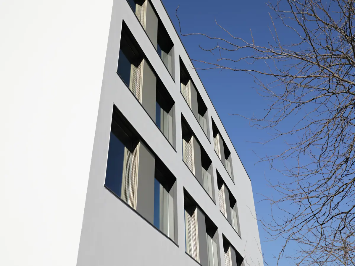 Nahaufnahme einer Gebäudefassade mit großen Fenstern und beige-farbenen Verschattungen, aufgenommen aus einem Winkel gegen den klaren blauen Himmel, mit Ästen im rechten Bereich des Bildes.