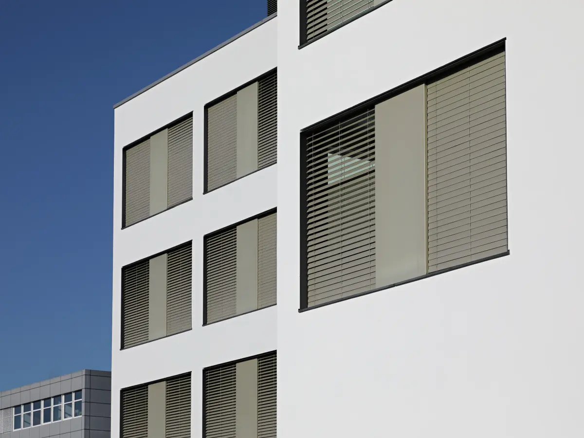 Detailaufnahme einer modernen, weißen Gebäudefassade mit großen Fenstern und beige-farbenen Jalousien, vor klarem, blauem Himmel.