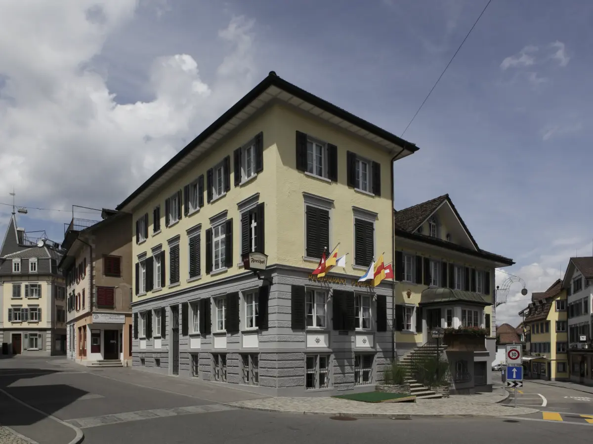 Historisches Eckgebäude mit gelber Fassade, schwarzen Fensterläden und Schweizer Flaggen an der Vorderseite.