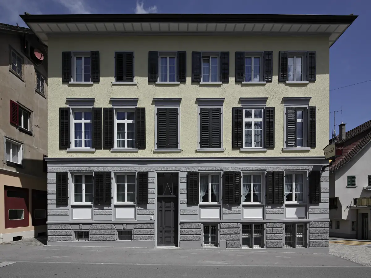 Fassade eines gelb-grauen Hauses mit schwarzen Fensterläden.