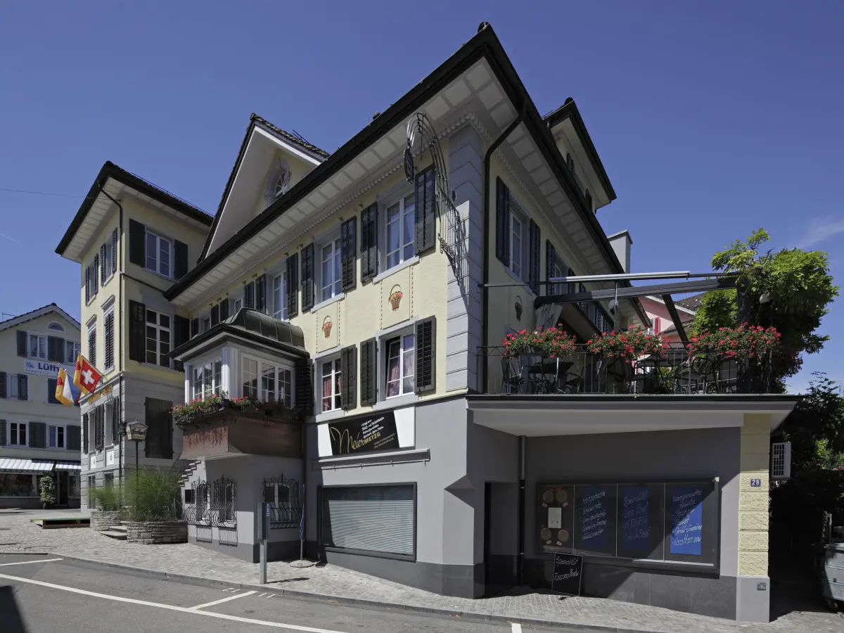 Gelb gestrichenes historisches Gebäude mit Balkon, schwarzen Fensterläden und Blumenschmuck, seitliche Ansicht.