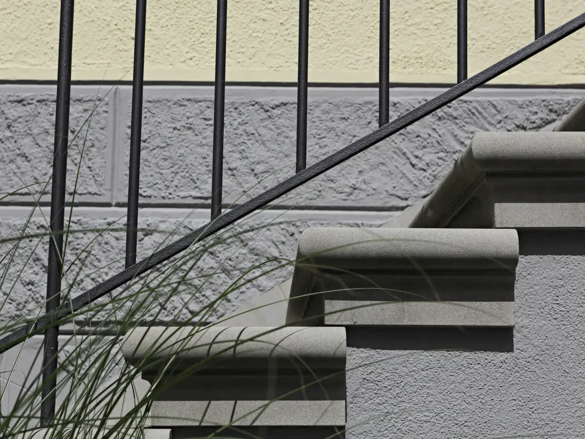 Detailaufnahme einer Steintreppe mit schwarzem Geländer und gelb-grauer Fassade im Hintergrund.