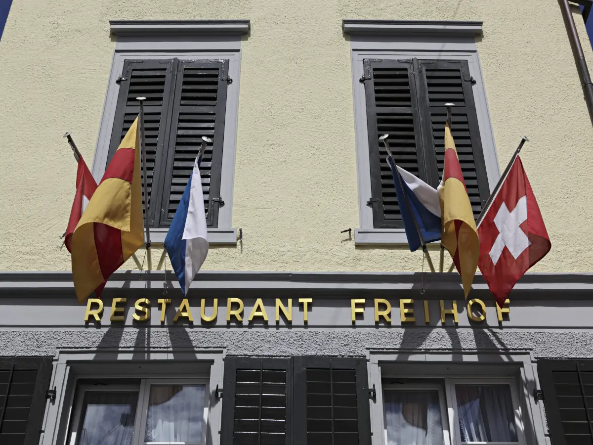 Fassade des Restaurants Freihof mit gelber Beschriftung und mehreren Flaggen, darunter die Schweizer Flagge.