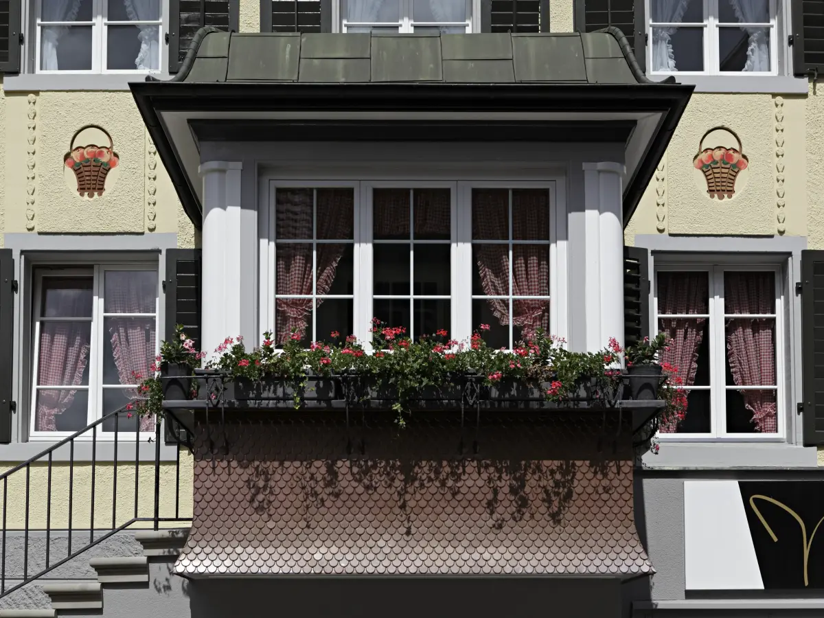 Detail eines traditionellen Erkers mit Blumenbepflanzung, großen Fenstern und gelber Fassade mit dekorativen Elementen.