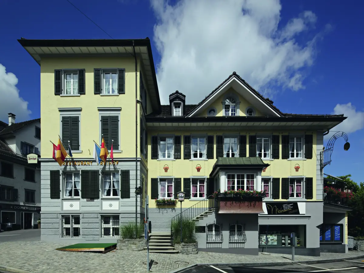 Gelb gestrichenes, historisches Gebäude mit schwarzen Fensterläden, Balkon und mehreren Eingängen.