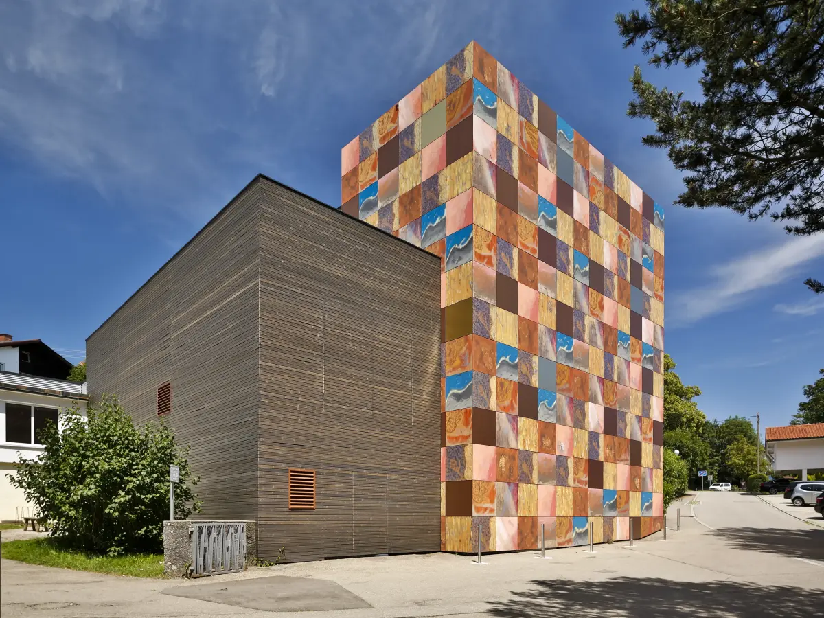 Edificio moderno con facciata a riquadri multicolori e parete laterale in legno scuro, lungo una strada sotto il cielo azzurro.