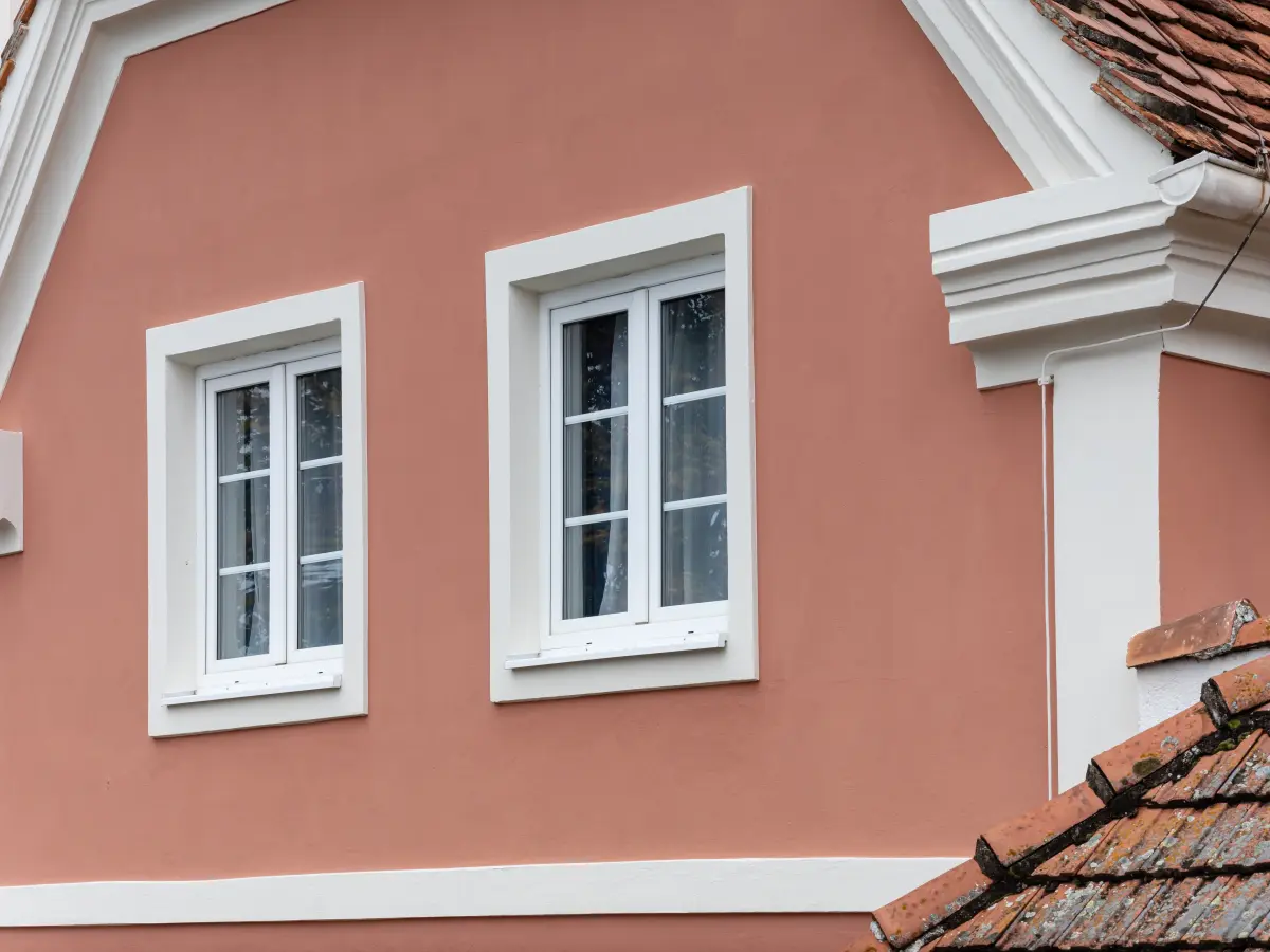 Zwei weiße Sprossenfenster mit dicker weißer Stuckumrandung in einer warmen, terrakottafarbenen Hausfassade.
