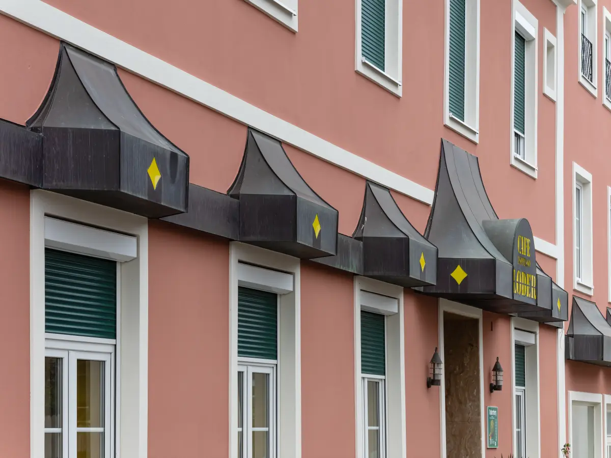 Terrakottafarbene Hausfassade mit dunklen, geschwungenen Vordächern, die mit gelben Rauten verziert sind, darunter Fenster mit grünen Rollläden.