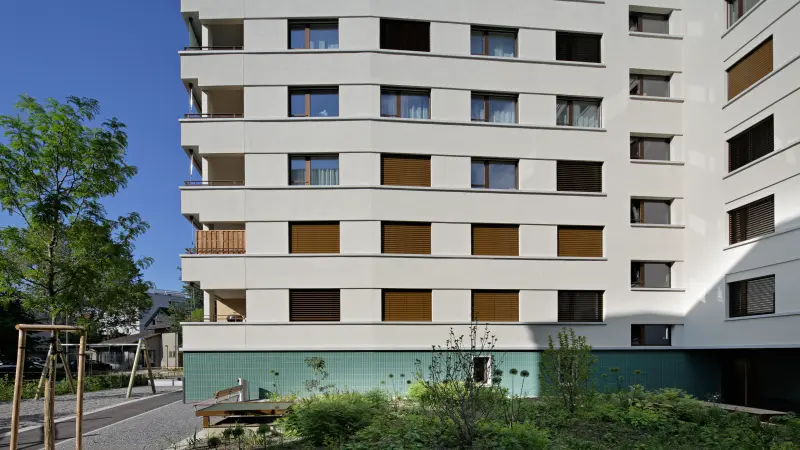 Wohngebäude mit modernen weißen Fassaden, braunen Fensterläden und grün gekacheltem Sockelbereich, umgeben von Bäumen und Grünflächen.