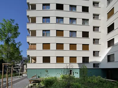 Wohngebäude mit modernen weißen Fassaden, braunen Fensterläden und grün gekacheltem Sockelbereich, umgeben von Bäumen und Grünflächen.