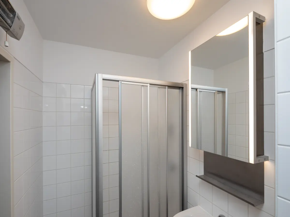 Modernes Badezimmer mit weiß gefliesten Wänden, einer Duschkabine mit Glastüren und einem beleuchteten Spiegel über dem Waschbecken.