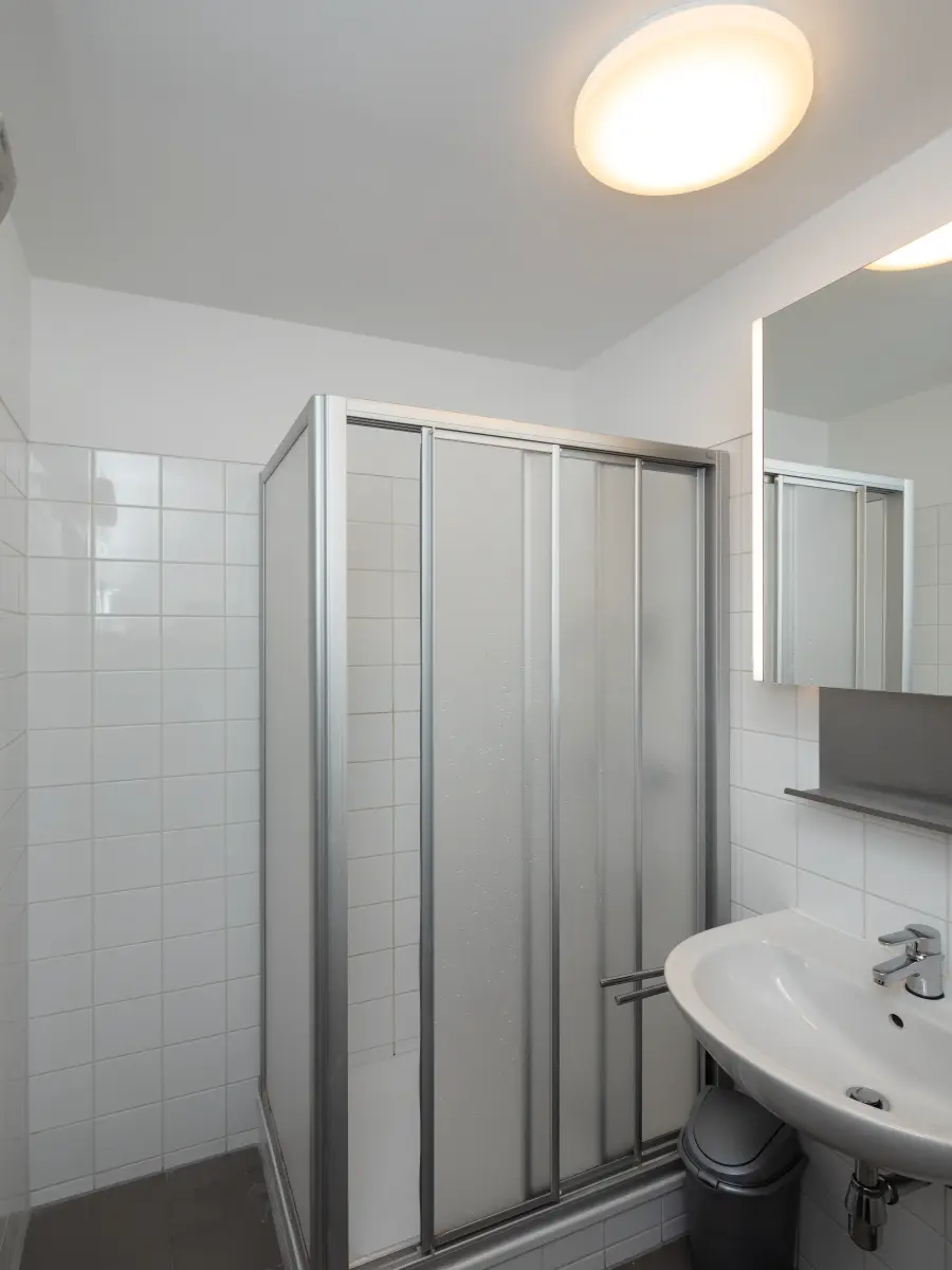 Kompaktes Badezimmer mit weiß gefliesten Wänden, einer Duschkabine mit mattierten Glastüren, einem Waschbecken und beleuchtetem Spiegel.