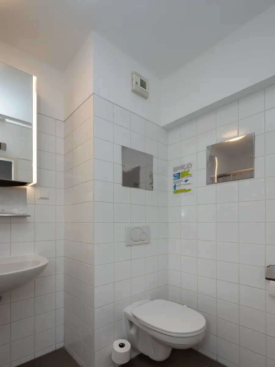Modernes Badezimmer mit weiß gefliesten Wänden, Wandtoilette mit Spülsystem, einem kleinen Waschbecken und einem beleuchteten Spiegel.