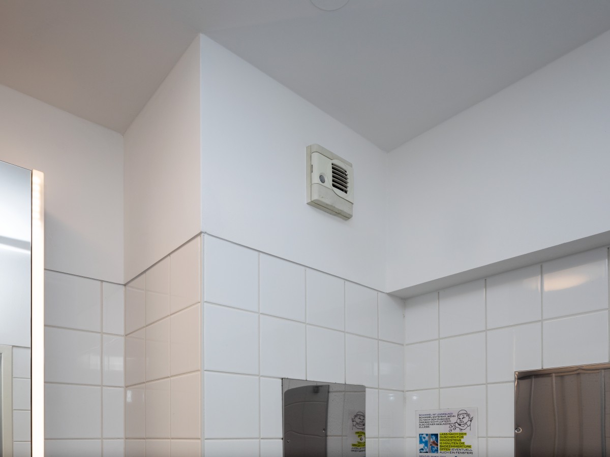 Detailansicht einer weiß gefliesten Badezimmerwand mit integriertem Lüftungssystem und Spiegeln.