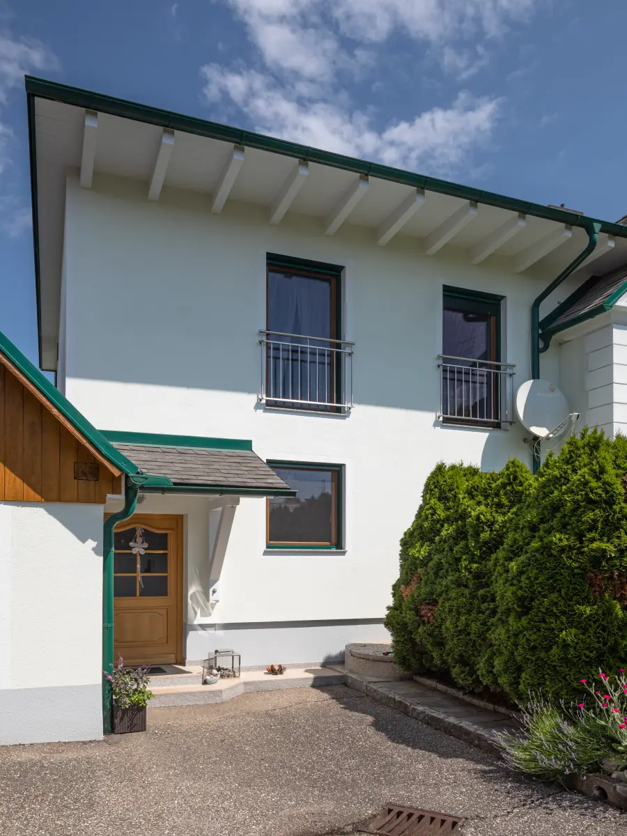 Weiße Hausfassade mit Holztür, Balkonen, dunklen Fensterrahmen und sichtbaren Dachsparren.