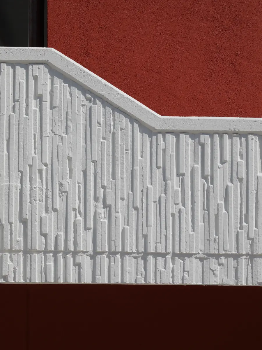 Detail einer roten Fassade mit strukturiertem weißem Beton-Relief und klaren geometrischen Linien.