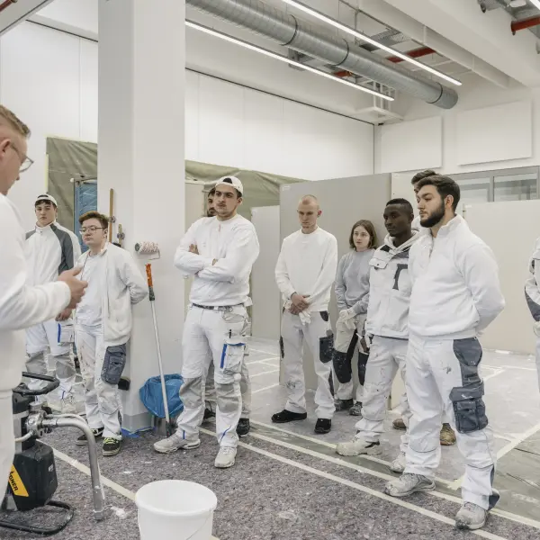 Eine Gruppe von Personen in Arbeitskleidung steht in einem hellen, industriellen Raum und hört einer Person zu, die etwas erklärt. Im Vordergrund sind Werkzeuge und ein Eimer sichtbar.
