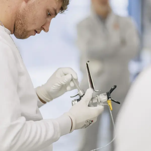 AI Eine Person in weißer Arbeitskleidung und Handschuhen inspiziert ein Farbspritzgerät, während im Hintergrund weitere Personen unscharf zu erkennen sind. Der Fokus liegt auf dem Gerät und den präzisen Handbewegungen.