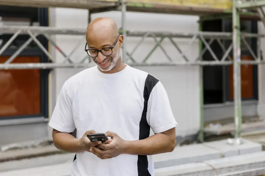  Eine Person steht vor einer Baustelle mit Gerüsten und hält ein Smartphone in den Händen. Im Hintergrund sind Fenster und Gebäudeteile sichtbar.