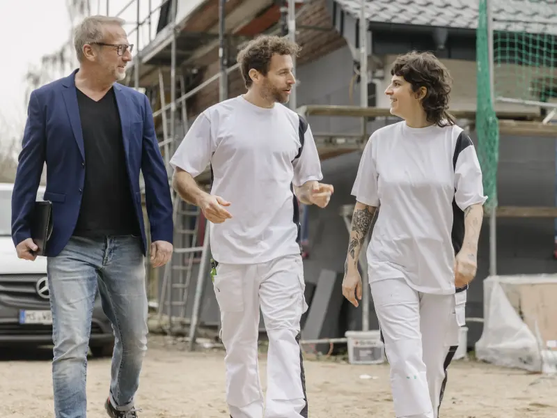 Bauleiter und zwei Maler unterhalten sich vor einem Gebäude mit Gerüst