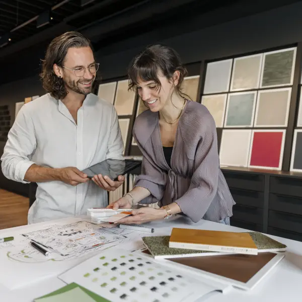 I Zwei Personen stehen an einem Tisch in einem modernen Showroom und betrachten Muster und Farbkarten. Auf dem Tisch liegen weitere Materialien, ein Katalog und Arbeitsunterlagen. Im Hintergrund sind Wandtafeln mit verschiedenen Oberflächenmustern ausgestellt.