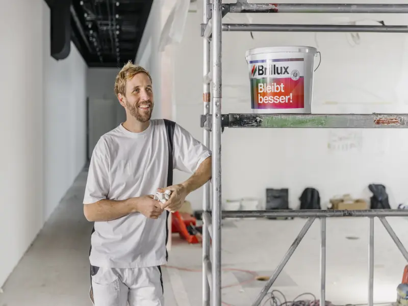 Ein Maler steht an einem Baugerüst auf dem Brillux Farbeimer stehen