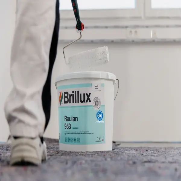 Maler mit Farbroller und Eimer Brillux Raulan 953 beim Streichen einer Innenwand.