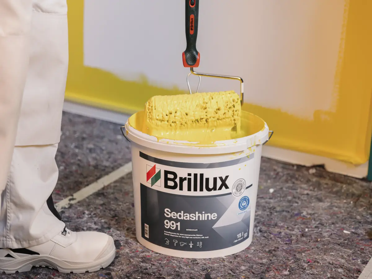 Geöffneter Brillux-Farbeimer mit gelber Sedashine 991, in den ein Maler einen Farbroller hält.