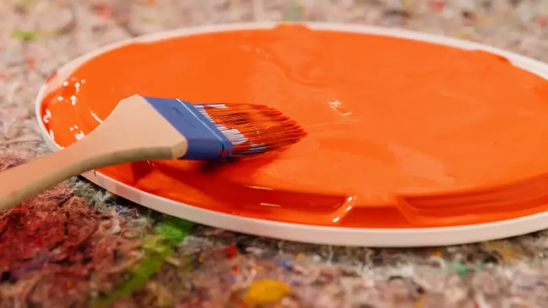 Deckel eines Farbeimers mit orangener Farbe darauf und einem Pinsel