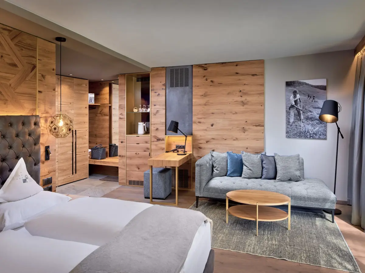 Camera d'albergo con finiture in stile legno, letto bianco e divano grigio.