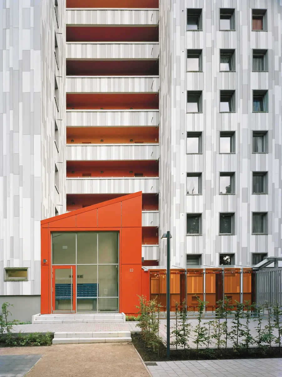 Eingangsbereich eines Hochhauses mit grauer Fassade und markanten orangefarbenen Elementen.