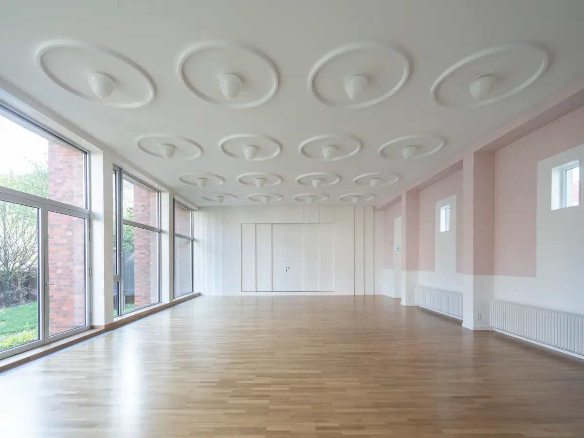 Heller, leerer Saal mit großen Fenstern, Holzboden und runden Deckenleuchten.