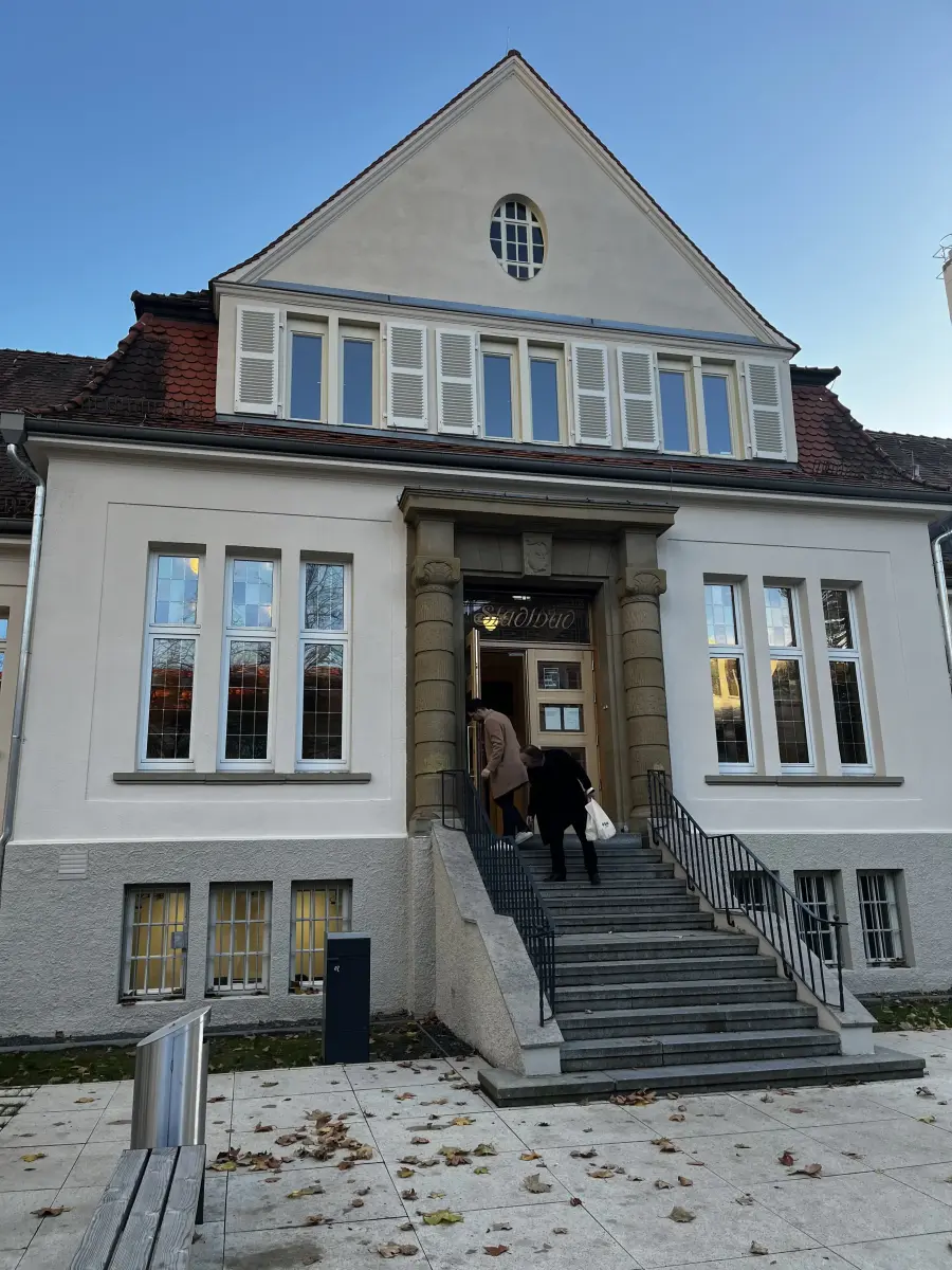 Helles, historisches Gebäude mit Giebeldach, das über eine Steintreppe von zwei Personen betreten wird.