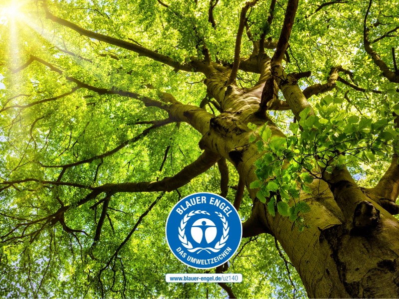 Ein grüner Baum im Sonnenlicht mit Blauem Engel Logo