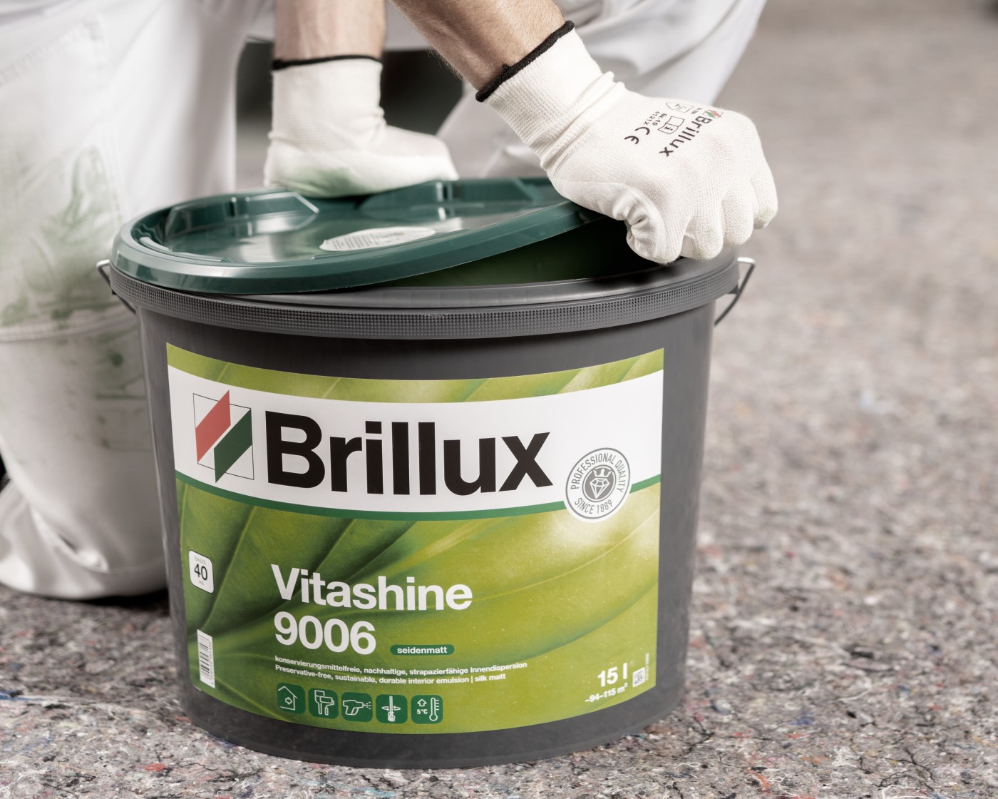 Ein Maler mit weißen Handschuhen öffnet einen großen, dunkelgrauen Eimer Brillux Vitashine 9006 Wandfarbe.