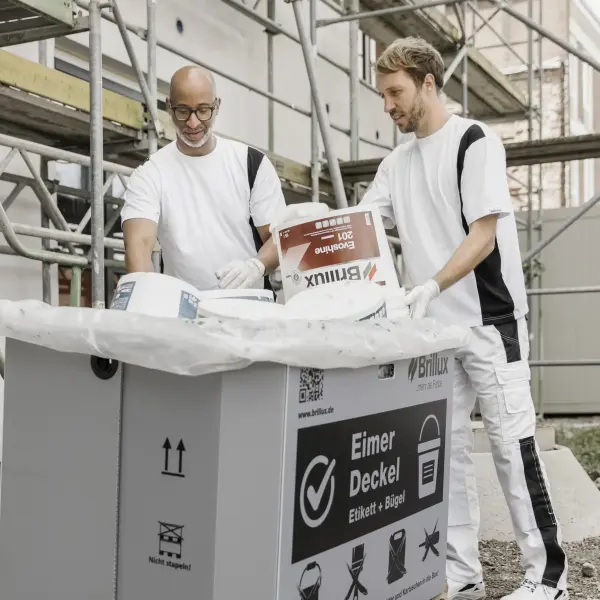 Zwei Maler werfen leere Brillux Farbeimer in einen Mülleimer auf der Baustelle
