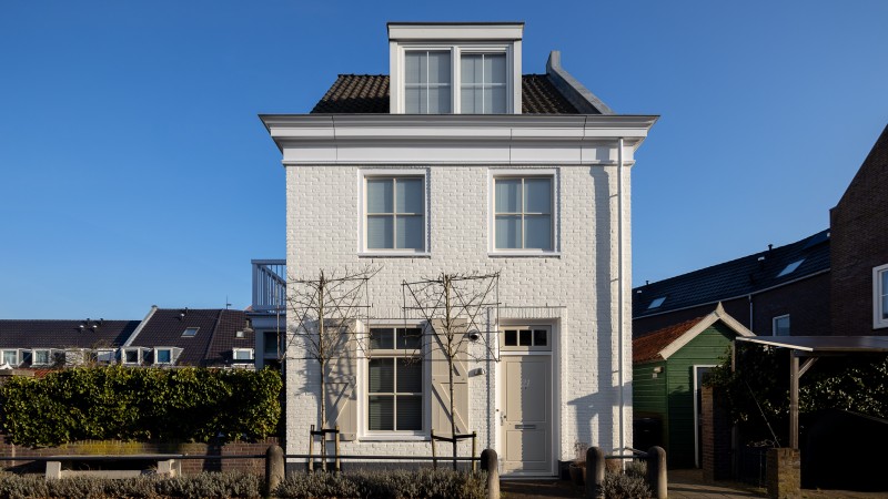 Ein einzelnes weißes Backsteinhaus mit zwei Stockwerken, einem Dachbodenfenster und einem grünen Gartenzaun davor, fotografiert bei blauem Himmel.