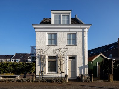 Ein einzelnes weißes Backsteinhaus mit zwei Stockwerken, einem Dachbodenfenster und einem grünen Gartenzaun davor, fotografiert bei blauem Himmel.