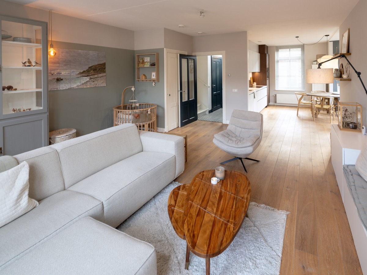 Ein moderner Wohnbereich mit einem weißen Sofa, einem Holztisch in Herzform, einem Kinderbett und einer offenen Küche im Hintergrund.