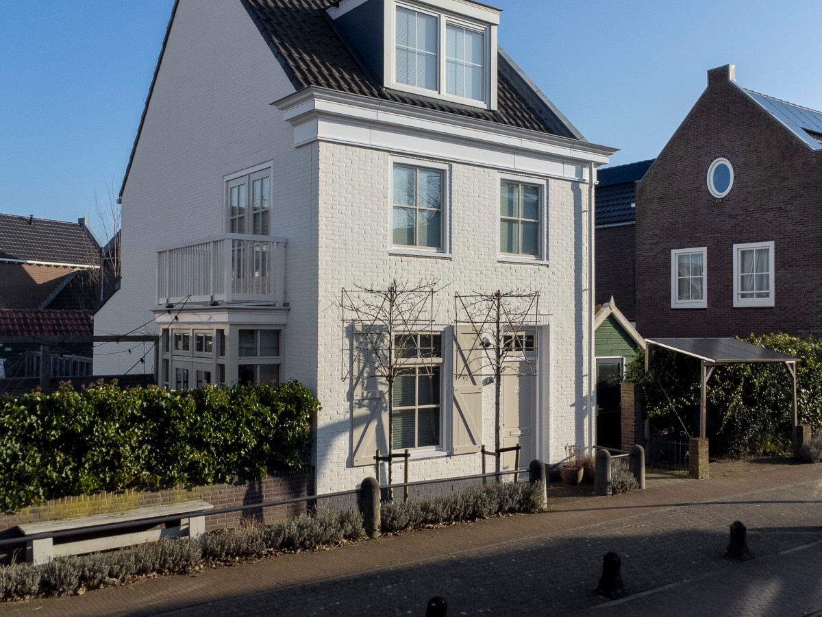 Zwei Häuser an einer Straße, eines weiß gestrichen mit Balkon und eines aus rotem Backstein, bei klarem Himmel.