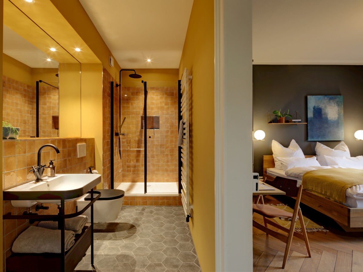 Modernes Hotelzimmer mit angrenzendem Bad, gelben Wänden, verglaster Dusche und gemütlichem Schlafzimmer mit Holzbett und Dekolampen.