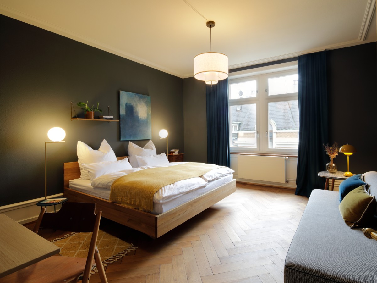 Stilvolles Hotelzimmer mit dunkel gestrichenen Wänden, Parkettboden, großem Fenster mit blauen Vorhängen sowie Sofa und kleinem Beistelltisch.