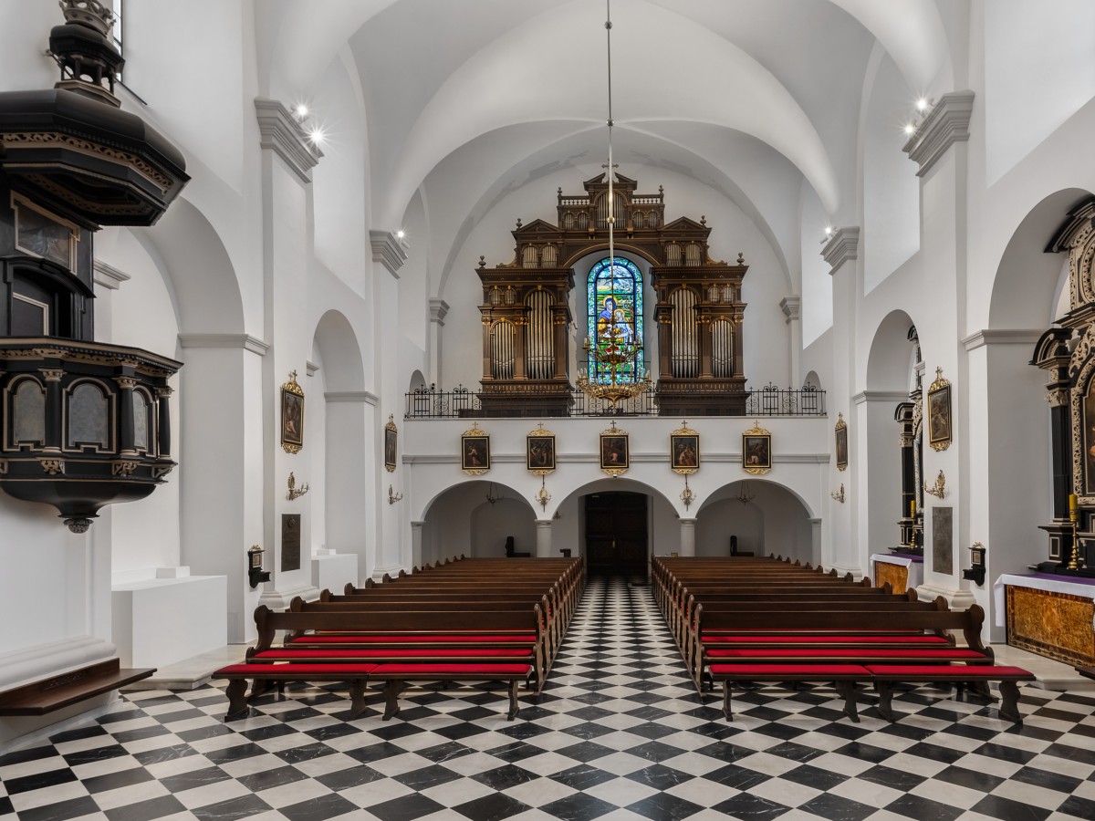 Eine große Kirche mit einem schwarz-weiß karierten Boden und Holzbänken, die zu einem verzierten Altar und einer Orgel am Ende des Ganges führen.