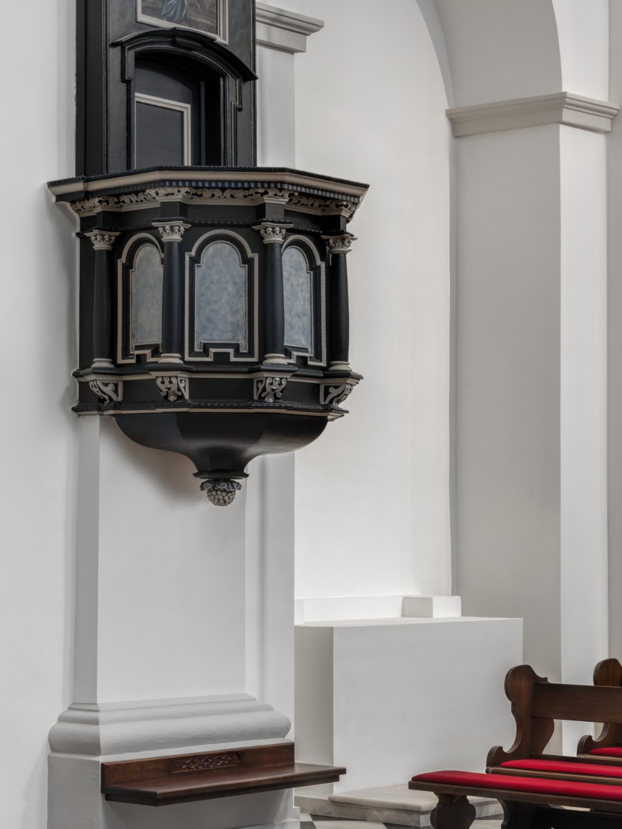 Eine schwarze, verzierte Kanzel an einer weißen Kirchenwand über einem schwarz-weiß gefliesten Boden.
